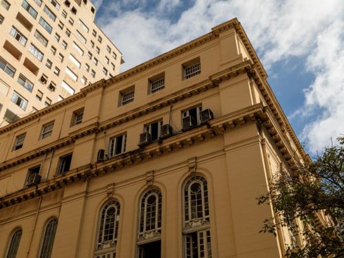 Edifício antigo de fachada bege, com janelas altas em arco e detalhes ornamentais. Algumas janelas possuem aparelhos de ar-condicionado. Ao fundo, aparece um prédio mais moderno e alto. O céu está azul com nuvens brancas, e há galhos de uma árvore no canto direito inferior.