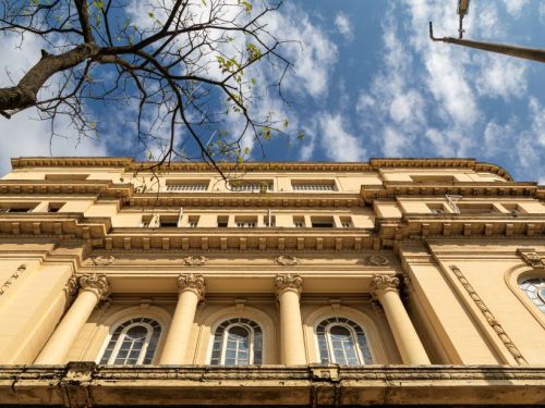 Uma foto tirada de baixo para cima de um prédio cor bege em estilo clássico. A imagem é dominada pela fachada do edifício, com colunas e janelas arqueadas. No canto superior esquerdo, um galho de árvore se estende para dentro do quadro, e no canto superior direito, um poste de luz vertical está visível. O céu está azul com nuvens brancas.