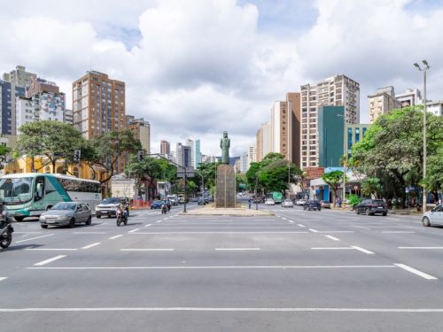 Uma vista ampla de um cruzamento movimentado em uma cidade. No centro da rua, sobre um pedestal escuro, ergue-se uma estátua de bronze de uma pessoa. Há vários carros, motos e um ônibus circulando nas pistas. Ao redor, diversos prédios altos e árvores verdes compõem a paisagem urbana, sob um céu com nuvens brancas.