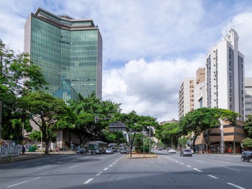 Uma rua larga e movimentada no centro de uma cidade. Há vários carros e um ônibus passando. Nas laterais da rua, há muitas árvores frondosas e edifícios altos. À esquerda, destaca-se um prédio moderno com fachada de vidro verde. À direita, outro prédio alto de cor clara. O céu está parcialmente nublado.