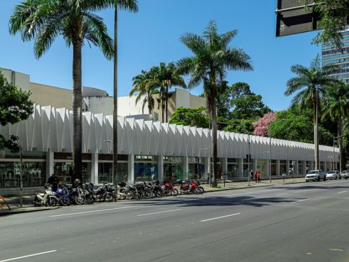 Uma rua larga com asfalto escuro e faixas brancas se estende em primeiro plano. À esquerda, na calçada, várias motocicletas estão estacionadas. Do lado direito, alguns carros estão parados. Ao longo da rua, há um longo prédio moderno de cor branca, o Palácio das Artes, com uma fachada que possui um padrão geométrico e grandes janelas no térreo, que parecem ser vitrines. Palmeiras altas e árvores verdes, uma delas com flores cor-de-rosa, enfeitam o ambiente sob um céu azul claro.