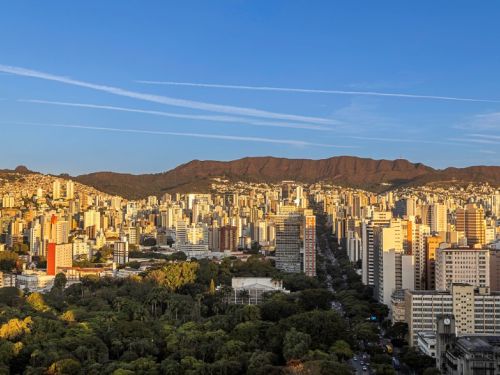 Uma vista panorâmica de uma cidade sob um céu azul claro. A luz dourada do sol de fim de tarde ilumina muitos prédios altos e claros. Em primeiro plano, uma grande área verde densa, como um parque, se estende. Ao fundo, uma cadeia de montanhas escuras se eleva, e uma longa avenida arborizada atravessa a cidade, com carros pequenos visíveis.