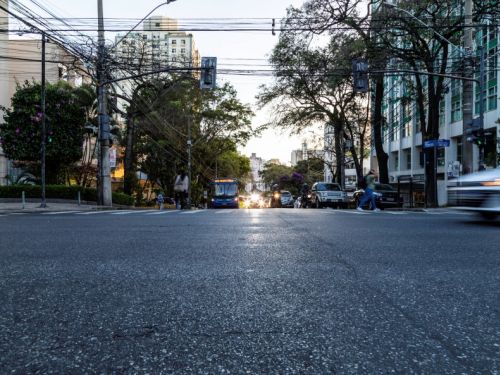 Uma rua de cidade grande, vista de perto do chão, olhando para frente em direção ao cruzamento. No centro, há um ônibus azul vindo em direção à câmera, cercado por carros com faróis acesos. À direita, um carro branco passa rápido, deixando efeito de movimento borrado. Em ambos os lados da rua há prédios altos, árvores grandes e muitos fios elétricos aéreos cruzando o espaço. Algumas pessoas atravessam a rua a pé. O céu está claro, provavelmente no fim da tarde, com luz suave. A cena transmite o movimento co