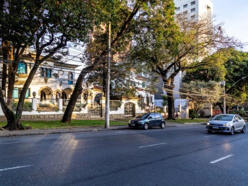 Uma rua arborizada, com casas antigas e árvores de grande porte. Um carro cinza está estacionado à direita da via, e um outro carro prateado trafega na mesma direção, mais próximo ao observador. As casas do lado esquerdo da rua têm fachadas em estilo colonial, com varandas e grades brancas. Ao fundo, um prédio de apartamentos mais moderno contrasta com as construções históricas.