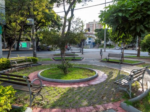 Uma pequena praça urbana, decorada com jardins simples e bancos de ferro com assentos de madeira. No centro há um canteiro redondo com grama e uma pequena árvore, cercado por uma borda de cimento e caminho de tijolos vermelhos. Outros canteiros e árvores menores se espalham pelo espaço, criando um ambiente agradável e sombreado. A praça fica em uma esquina de uma avenida, e ao fundo vê-se prédios residenciais, postes com muitos fios e semáforos. Algumas pessoas circulam de bicicleta ou moto ao longe.