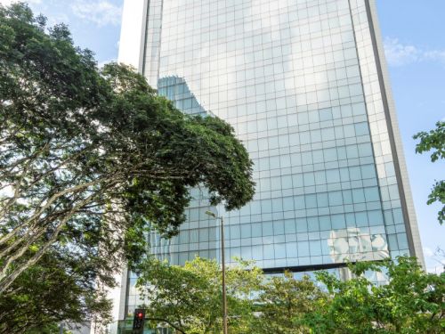 Um prédio muito alto e moderno, de fachada espelhada em tons claros, refletindo o céu azul com nuvens e parte das construções vizinhas. No topo, há o logotipo de uma empresa em letras laranja. Algumas pequenas árvores estão plantadas em um espaço elevado na própria lateral do edifício, dando destaque verde em meio ao vidro. Na frente, há uma avenida com carros estacionados e outros circulando, além de semáforos acesos em vermelho. Árvores grandes ladeiam a via, misturando natureza com a paisagem urbana vert