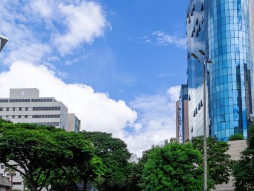 Um cruzamento urbano em dia ensolarado. Em destaque, um prédio moderno de vidro azul à direita, contrastando com árvores grandes e outros edifícios ao fundo. Carros atravessam a avenida, e o céu azul com nuvens brancas completa a cena, trazendo leveza ao ambiente movimentado da cidade.