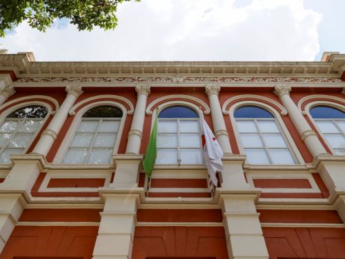 A imagem mostra a fachada de um prédio histórico de cor vermelha com detalhes em branco, janelas altas em arco e ornamentos decorativos na parte superior. Duas bandeiras, uma verde e outra branca, estão hasteadas no centro da construção.