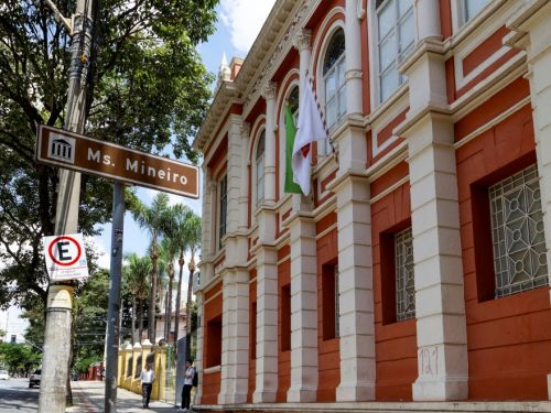A imagem mostra a fachada lateral de um prédio histórico vermelho com detalhes em branco, janelas grandes em arco e bandeiras hasteadas. Em frente, há uma placa indicando “Ms. Mineiro” e pessoas caminhando pela calçada de pedra decorada.