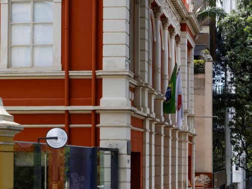 A imagem mostra a fachada lateral de um prédio histórico em tom vermelho-alaranjado com detalhes em branco, janelas altas em arco e bandeiras hasteadas. Na calçada de pedra, algumas pessoas caminham em frente à entrada com porta de vidro.