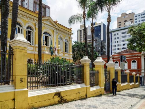 A imagem mostra dois prédios históricos cercados por um muro e grade de ferro: à esquerda, um casarão amarelo com detalhes brancos, janelas altas e bandeiras na fachada; à direita, um edifício vermelho com colunas e ornamentos claros. Há palmeiras no jardim e pessoas caminhando na calçada de pedra em frente. Ao fundo, aparecem prédios modernos.