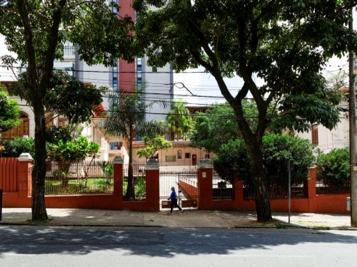 Prédio histórico de dois andares em tons claros, cercado por um muro vermelho com grades de ferro. Na frente há um jardim com árvores e palmeiras, além de grandes árvores na calçada que projetam sombra sobre a rua. Uma pessoa atravessa o portão de entrada.