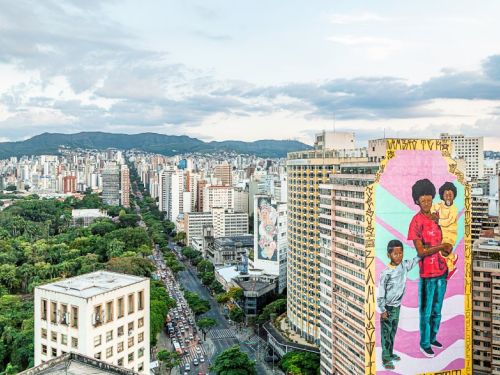 Uma vista aérea panorâmica de uma cidade movimentada com muitos prédios altos e uma longa avenida arborizada cheia de carros. Ao fundo, há montanhas sob um céu claro com nuvens. No lado direito da imagem, um edifício exibe um grande e colorido mural: ele retrata uma mulher de pele escura segurando um bebê, e ao seu lado, um menino maior, em um fundo de tons de rosa e azul claro.