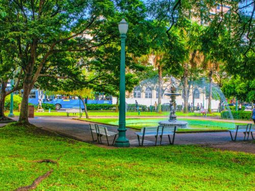 Fonte circular com escultura central de figuras mitológicas, cercada por jatos d’água arqueados. Ao redor, bancos de ferro, postes verdes de luminária clássica e árvores frondosas que criam sombra. Grama bem cuidada cobre o chão, enquanto ao fundo aparecem palmeiras, prédios históricos e pedestres em movimento.