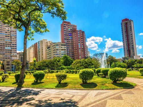 A fonte central da praça está em primeiro plano, com vários jatos de água esguichando para o alto a partir de um grande tanque octogonal de cor azul claro. Ao fundo, do outro lado da avenida, erguem-se edifícios altos de arquitetura clássica, em tons terrosos, sob um céu azul com nuvens.