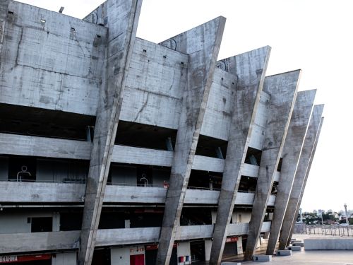 A imagem mostra uma grande seção da fachada do estádio Mineirão, a arquitetura de concreto, com seus pilares inclinados e robustos, é o destaque, revelando a passagem do tempo através das texturas e marcas. A luz do dia ilumina o edifício de forma uniforme, e é possível ver algumas pessoas nas sacadas e a paisagem da cidade ao fundo. O ponto de vista, de baixo para cima, enfatiza a escala imponente da construção.