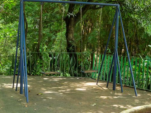 Estrutura metálica azul de balanço com duas correntes e assentos suspensos, instalada sobre piso cimentado. Ao fundo, há árvores altas de folhagem densa e vegetação variada.