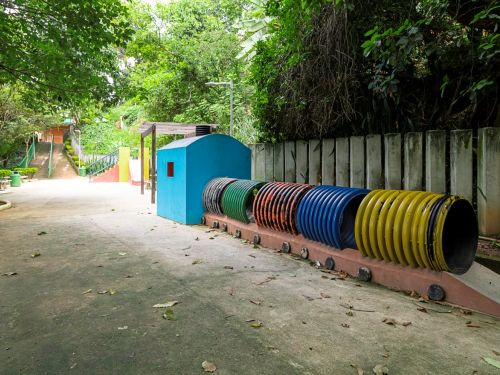 Brinquedo infantil com túneis de tubos plásticos coloridos em amarelo, azul, laranja e verde. Estrutura apoiada em base de concreto no chão cimentado. Ao fundo, árvores e mureta de madeira clara delimitam o espaço.