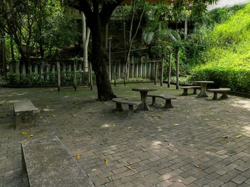 Praça pavimentada com blocos de concreto. No centro, mesas redondas e bancos circulares de cimento. Ao fundo, um banco comprido de concreto e brinquedos de madeira. Árvores grandes fazem sombra sobre a área.