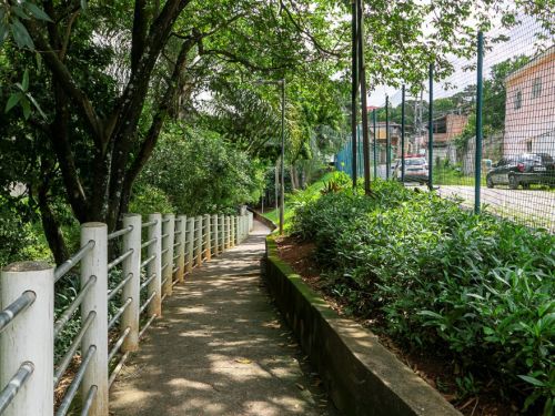 Caminho de cimento cercado por grades metálicas brancas à esquerda e canteiro elevado com arbustos à direita. Árvores grandes sombreiam o trajeto. Ao fundo, há uma quadra cercada por alambrado e casas visíveis.