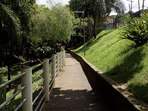 Caminho de cimento com corrimão branco à esquerda e barranco gramado à direita. Árvores altas projetam sombra no trajeto. No fundo, postes de iluminação e parte de uma quadra cercada por alambrado.