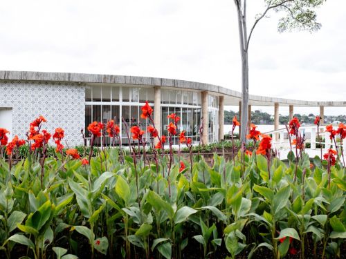 a Casa do Baile vista de um jardim florido. No primeiro plano, há um canteiro com muitas flores vermelhas e laranjas e folhas verdes. Atrás, a construção de forma circular tem grandes janelas de vidro e uma parede com azulejos azuis e brancos. Um telhado curvo, sustentado por colunas, se estende para o lado, com vista para a Lagoa da Pampulha.