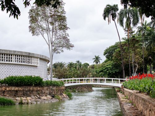 Lateral da Casa do Baile, com sua fachada arredondada e azulejos claros. Uma ponte branca e em arco atravessa um canal de água esverdeada. As margens são de pedra e ladeadas por plantas verdes, com um canteiro de flores vermelhas à direita. Ao fundo, uma densa linha de árvores, incluindo palmeiras, sob um céu nublado.