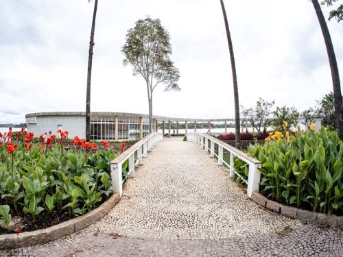 Vista frontal da Casa do Baile. Um caminho de pedras claras leva a uma ponte branca e curva, que é ladeada por jardins com flores vermelhas e amarelas. Ao fundo, a construção de forma circular tem grandes janelas de vidro. Ela está sobre a Lagoa da Pampulha, sob um céu com nuvens brancas e cinzas.