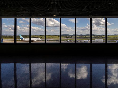 Através de uma ampla janela de vidro dividida em painéis verticais, vê-se a pista do aeroporto. Um avião branco com detalhes em azul está parado. Ao fundo, uma paisagem de campos verdes e morros sob um céu azul com nuvens. O piso polido do saguão reflete a imagem da janela.