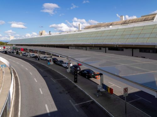 Vista elevada da área de embarque e desembarque do aeroporto. Uma longa cobertura translúcida se estende sobre a via onde carros estão parados em fila. À direita, a fachada do aeroporto com uma parede de vidro inclinada. O céu está azul com algumas nuvens brancas e o dia está ensolarado.