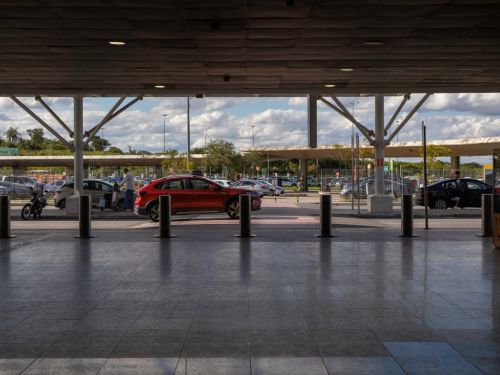 Vista de dentro do terminal, olhando para fora em direção à área de estacionamento e vias de acesso. A imagem é enquadrada por pilares e pelo teto do saguão, que estão em sombra. Ao fundo, carros, incluindo um SUV vermelho, e pessoas se movimentam sob um céu parcialmente nublado.