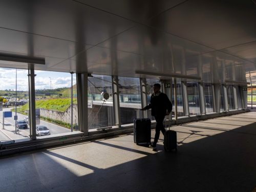 Um passageiro caminha por uma passarela envidraçada, puxando duas malas de rodinha. Ele está em silhueta contra a luz que entra pelas janelas. A passarela, com piso escuro e teto claro, projeta sombras geométricas no chão. Pela janela, vê-se a área externa com estacionamento e vegetação.