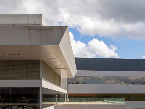 Detalhe da arquitetura externa do aeroporto, mostrando o encontro de diferentes blocos e materiais. Uma grande marquise de metal branco com linhas angulares se projeta sobre uma fachada de vidro. Ao fundo, outro bloco do edifício com revestimento cinza, sob um céu azul com nuvens.