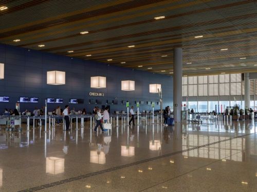 Amplo salão de check-in do aeroporto, com piso brilhante que reflete as luzes. Uma longa parede azul ao fundo tem a inscrição "CHECK-IN 3". Em frente, balcões de atendimento e filas de passageiros com carrinhos de bagagem. Grandes luminárias quadradas pendem do teto de ripas de madeira.
