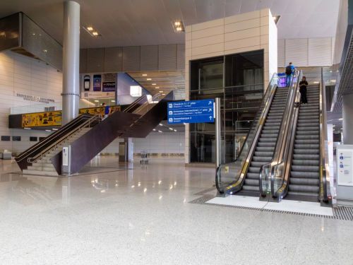 Saguão espaçoso e moderno do aeroporto, com piso de granito claro e brilhante. À direita, duas escadas rolantes em funcionamento. À esquerda, uma escada fixa de degraus de granito e corrimão de metal. Uma placa azul indica as direções para check-in, embarque e desembarque.