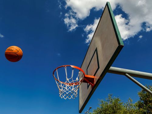 Uma foto tirada de baixo para cima, de uma cesta de basquete. Uma bola laranja está no ar, prestes a cair na cesta. A cesta, no lado direito, tem um aro laranja e uma tabela branca. O céu é azul e claro, com nuvens brancas e esparsas.