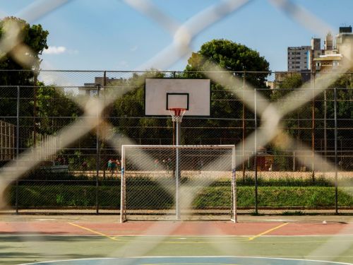 Uma quadra esportiva, vista através de uma cerca de arame em primeiro plano. A quadra tem o chão em tons de verde e vermelho, com um gol de futebol e uma cesta de basquete no centro. O local é cercado por árvores, com prédios altos visíveis ao fundo, sob um céu azul com nuvens brancas.