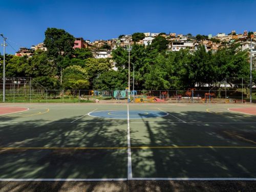 Uma quadra esportiva em um parque. O chão tem marcações em várias cores, como verde e azul. Atrás da quadra, um morro íngreme é coberto por uma vasta área de casas de diversas cores, que se estendem até o topo. O morro é separado da quadra por uma faixa de árvores verdes. O céu está azul e sem nuvens, com o sol projetando sombras no chão.