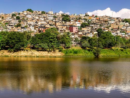 Um morro densamente ocupado por casas. O primeiro plano é um lago de água calma, que reflete a paisagem. Atrás do lago, uma fileira de árvores verdes separa a água da encosta. O morro íngreme é coberto por uma vasta área de casas de diversas cores, que se estendem até o topo. O céu é azul com nuvens brancas.