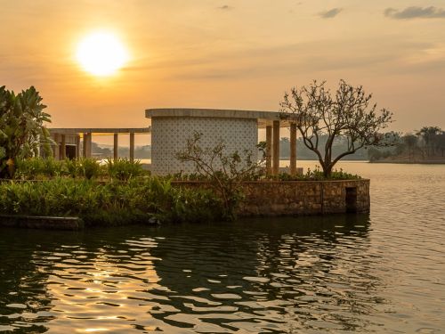 Casa do Baile na Lagoa da Pampulha, ao pôr do sol. O sol está no canto superior esquerdo e sua luz dourada se reflete na água, criando um brilho intenso e ondulado. A construção, em uma pequena ilha com vegetação, tem uma forma arredondada. Uma árvore de galhos finos e escuros se destaca, criando uma silhueta à direita do prédio.