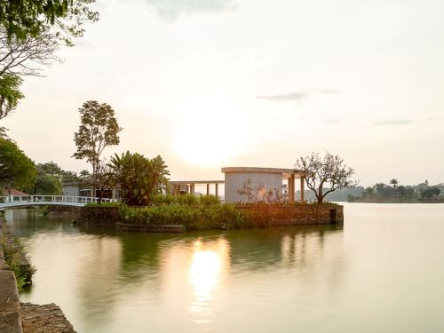 Casa do Baile ao pôr do sol na Lagoa da Pampulha. O lago está calmo e reflete a luz dourada do sol, que se esconde atrás da construção. A casa é uma estrutura circular, em uma pequena ilha, conectada à margem por uma ponte branca. No horizonte, há uma linha de árvores e prédios.