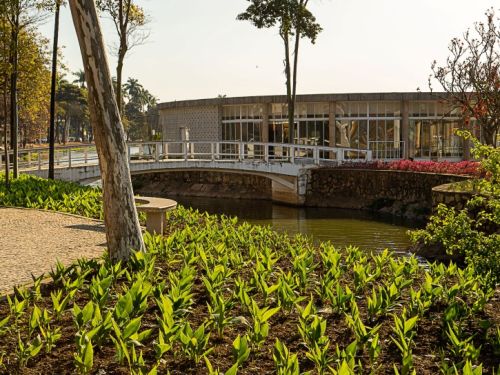 A Casa do Baile em um dia claro. Em primeiro plano, há um canteiro de plantas pequenas e verdes. Um pequeno canal de água e uma ponte branca levam até a construção, que é uma estrutura baixa e longa com grandes janelas de vidro. Árvores altas estão plantadas ao redor do local, sob um céu azul.