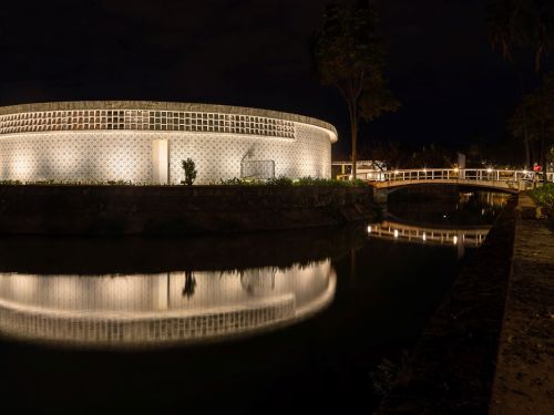 Casa do Baile à noite, totalmente iluminada. A construção circular tem a fachada com azulejos claros e padrões geométricos que brilham na escuridão. A luz do prédio se reflete de forma quase perfeita na água escura. Uma ponte branca e curva, também iluminada, liga a construção à margem.
