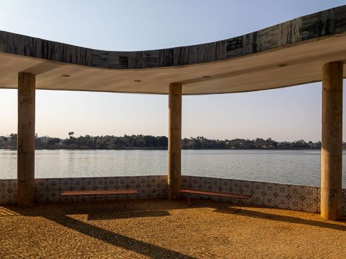 Uma vista da área externa Casa do Baile, de frente para a lagoa. O espaço coberto tem chão de pedras claras e é sustentado por colunas redondas. Uma mureta baixa com azulejos azuis e brancos circunda o local. Dois bancos de madeira estão posicionados na frente. A luz do sol da tarde projeta longas sombras no chão e, ao fundo, está a grande lagoa da Pampulha.