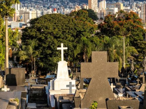 Um cemitério em Belo Horizonte, com a cidade e as montanhas ao fundo, sob um céu claro de fim de tarde. Em primeiro plano, uma variedade de túmulos preenche o espaço. No centro, destaca-se um túmulo branco brilhante com uma grande cruz branca no topo, contrastando com os túmulos escuros ao seu redor. Atrás dos túmulos, há uma linha de árvores verdes escuras, incluindo algumas palmeiras.