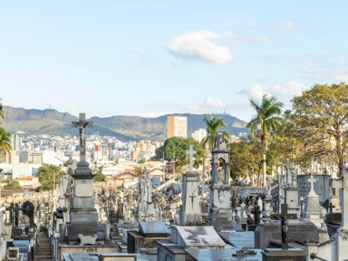 Um cemitério em uma colina, com uma vista da paisagem urbana e das montanhas ao fundo. Os túmulos, feitos de mármore e granito, estão dispostos em fileiras, preenchendo o primeiro plano da imagem. No plano intermediário e ao fundo, a cidade de Belo Horizonte é visível com seus edifícios e arranha-céus.