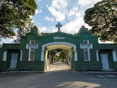 O portão de entrada de um cemitério no final de uma tarde. O portão é uma grande estrutura de alvenaria pintada de um tom de verde escuro, com um arco central que serve de entrada para veículos e pedestres. No topo, há uma cruz branca e uma inscrição que parece ser em latim. De cada lado do arco central, há duas janelas retangulares, cada uma com uma cruz branca vazada em sua grade.