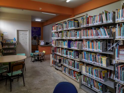 Sala de biblioteca com estantes metálicas cheias de livros organizados. Há mesas redondas com cadeiras escolares e um balcão de atendimento ao fundo. A iluminação é feita por lâmpadas fluorescentes.