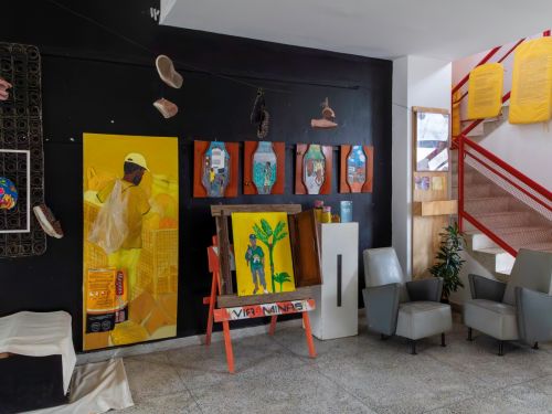Espaço expositivo com parede preta onde estão fixados objetos e quadros coloridos. No chão há duas poltronas cinza, e ao lado uma escada com corrimão vermelho. Obras incluem pinturas de pessoas e objetos.
