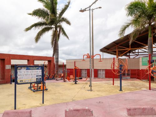 Área externa com equipamentos de academia ao ar livre em estrutura laranja e azul. No espaço há um piso de cimento pintado e um painel de instruções de exercícios. Palavras “Academia a Céu Aberto” aparecem na placa.